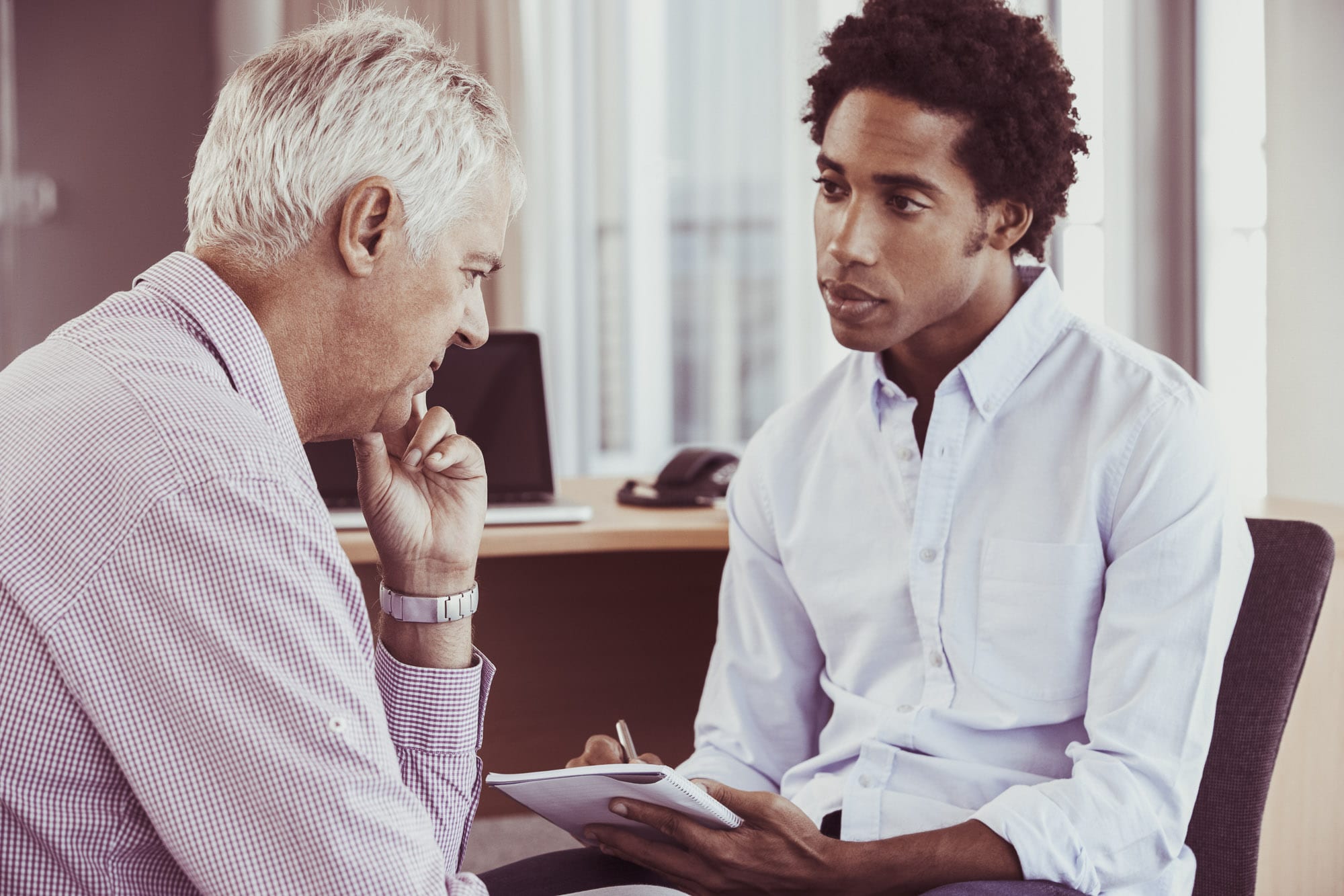 Does Mental Health Count as a Disability? A young black man interviewing an elderly white man. The elderly man looks deep in thought while the black man looks concerned. He appears to be taking notes with a notepad and pen in hand.